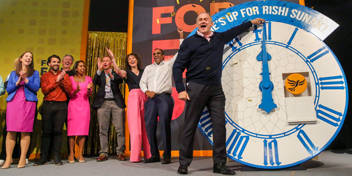 Ed Davey MP on stage at Autumn Conference with other Lib Dem figures and a giant clock labelled 'Time's up for Rishi Sunak!'
