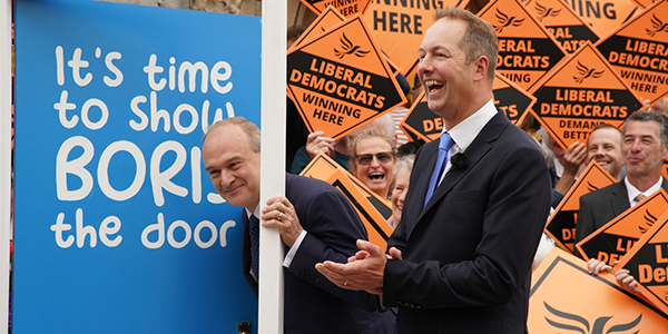 Ed Davey peers through a door which says 'It's time to show Boris the door' on it, next to Richard Foord celebrating his by-election win.