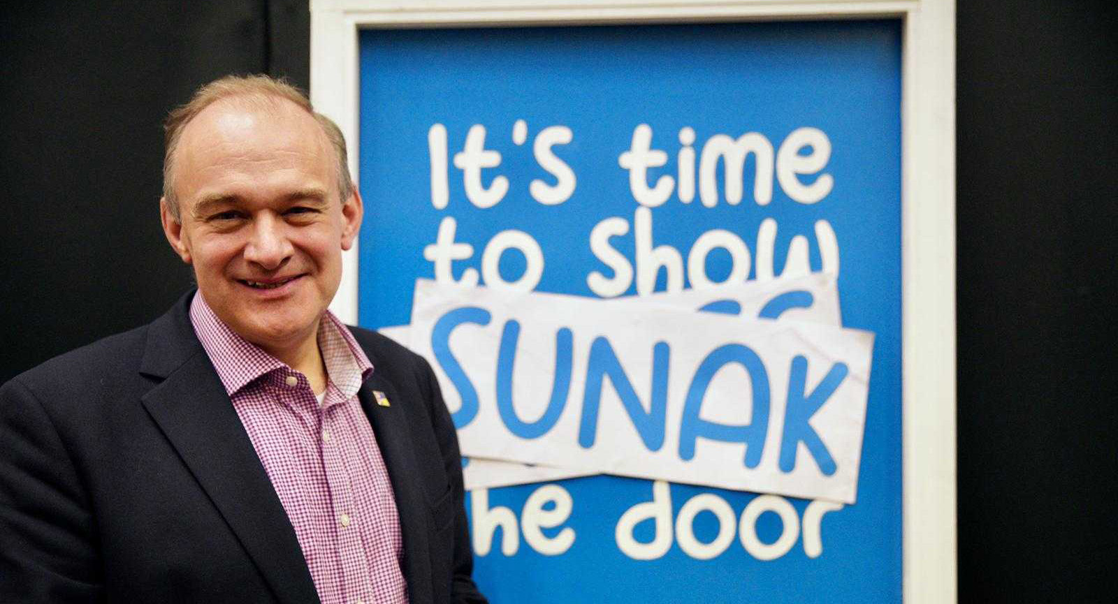 Ed Davey stood in front of a blue door that reads 'It's time to show Sunak the door'