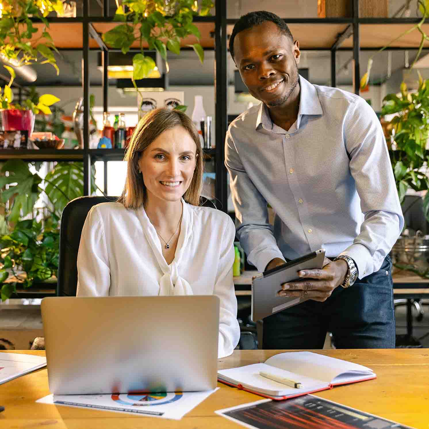Two people at a desk with one sitting down at a laptop and another standing and showing the other something on a tablet.