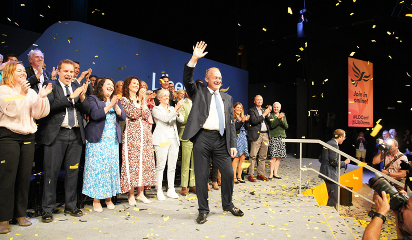 Ed Davey MP waves from the Conference stage with Lib Dem MPs standing behind him clapping and gold confetti falling.