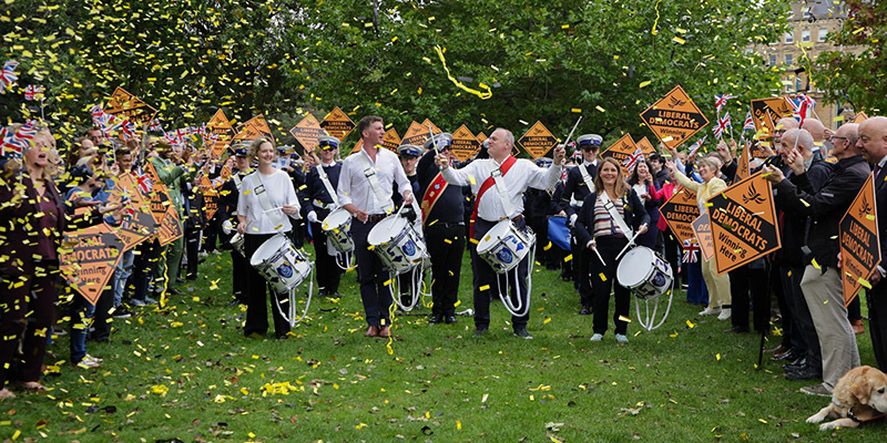 Anna Sabine, Ed Morello, Ed Davey, and Vikki Slade leading a marching band, flanked by Lib Dem supporters.