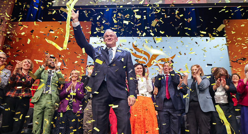 Ed Davey waving from the main stage with Lib Dem MPs applauding behind him.