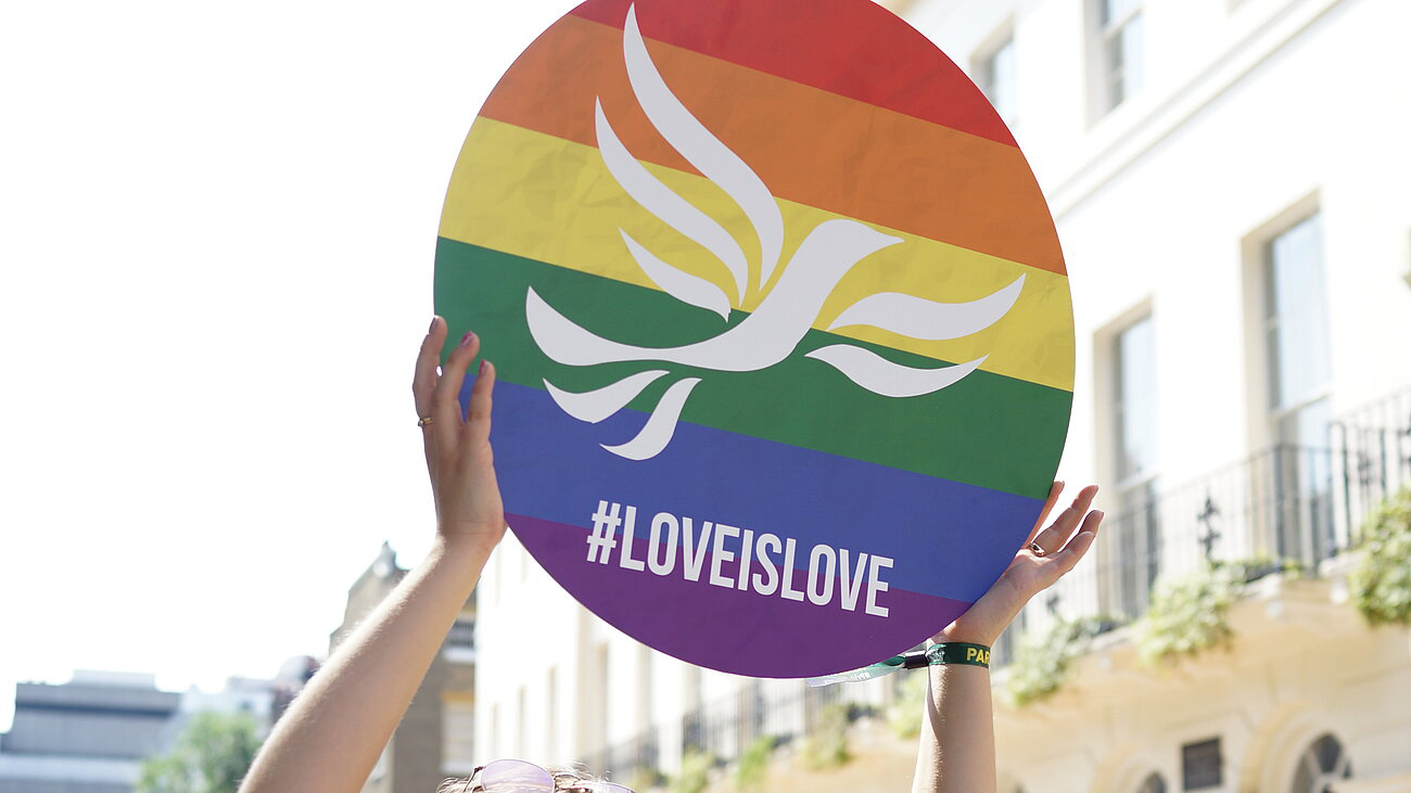 Someone holds up a circular banner with the Lib Dem logo on a rainbow flag background with the words #LoveisLove.