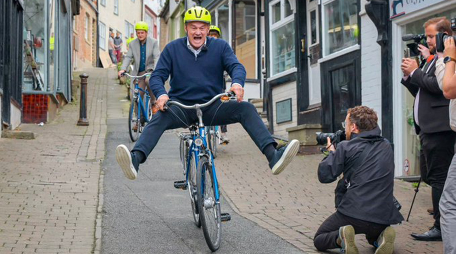 Ed Davey rides a bike down a steep street, lifting his feet off the peddles.