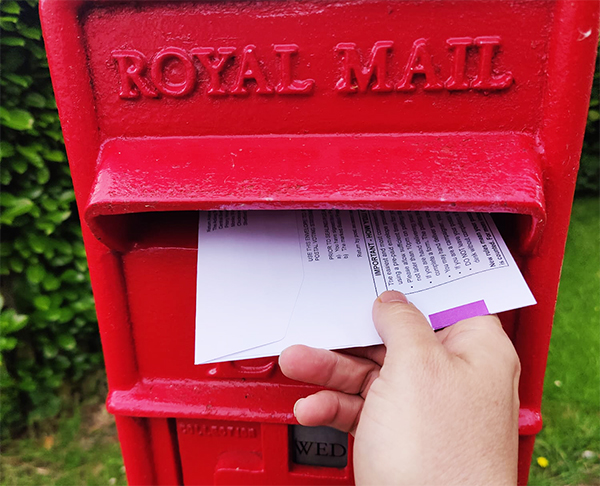 A red Royal Mail postbox, a hand is posting a postal vote for the General Election through it.