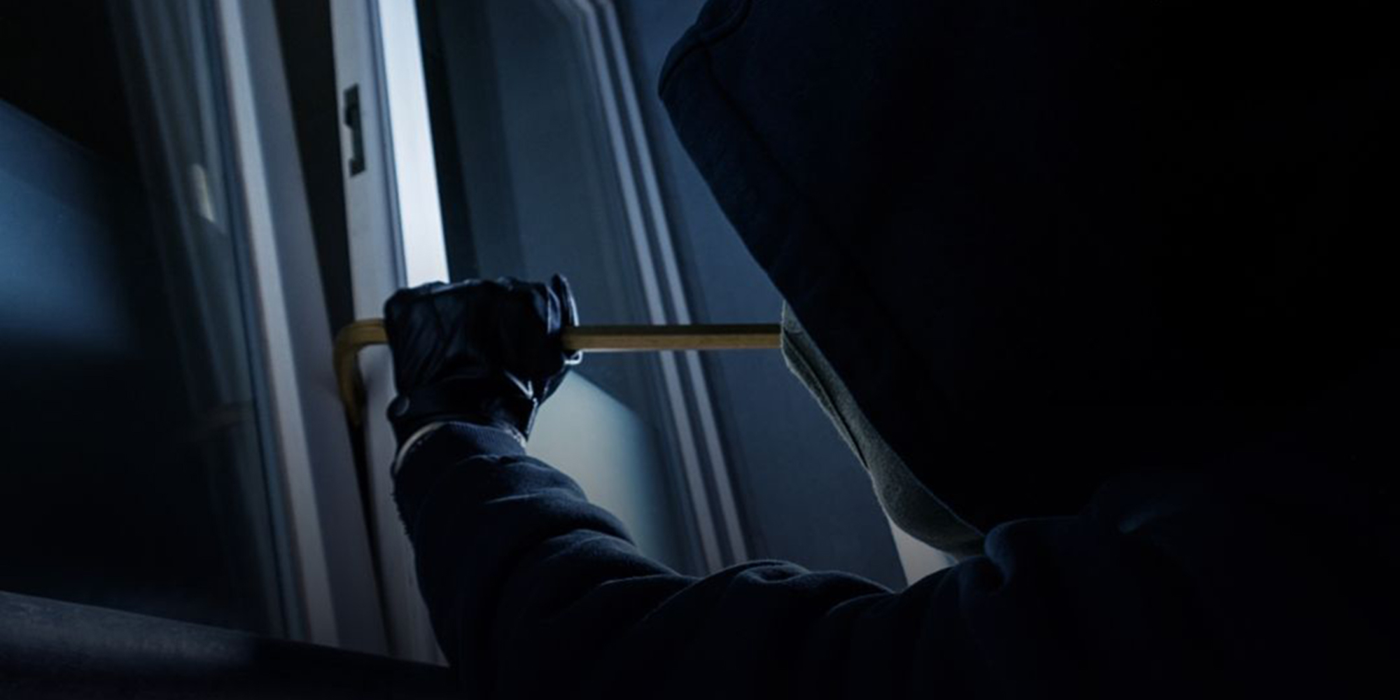 A burglar wearing a hood breaks through a window.